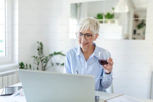 Beautiful middle aged businesswoman wearing rectangular glasses , sitting at home office with glass of wine on desk. Senior woman working from home