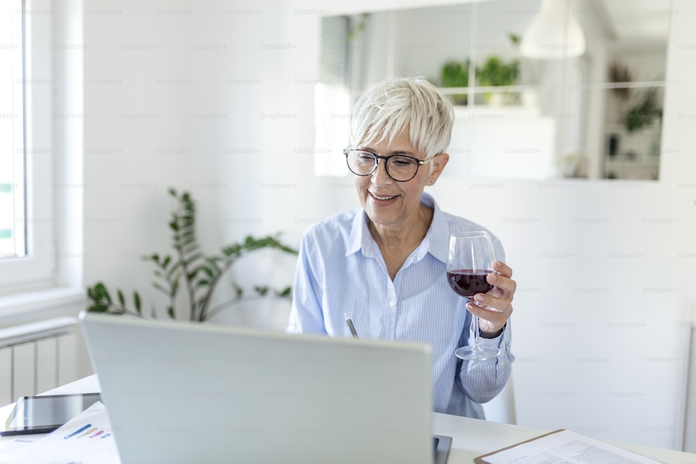 Beautiful middle aged businesswoman wearing rectangular glasses , sitting at home office with glass of wine on desk. Senior woman working from home