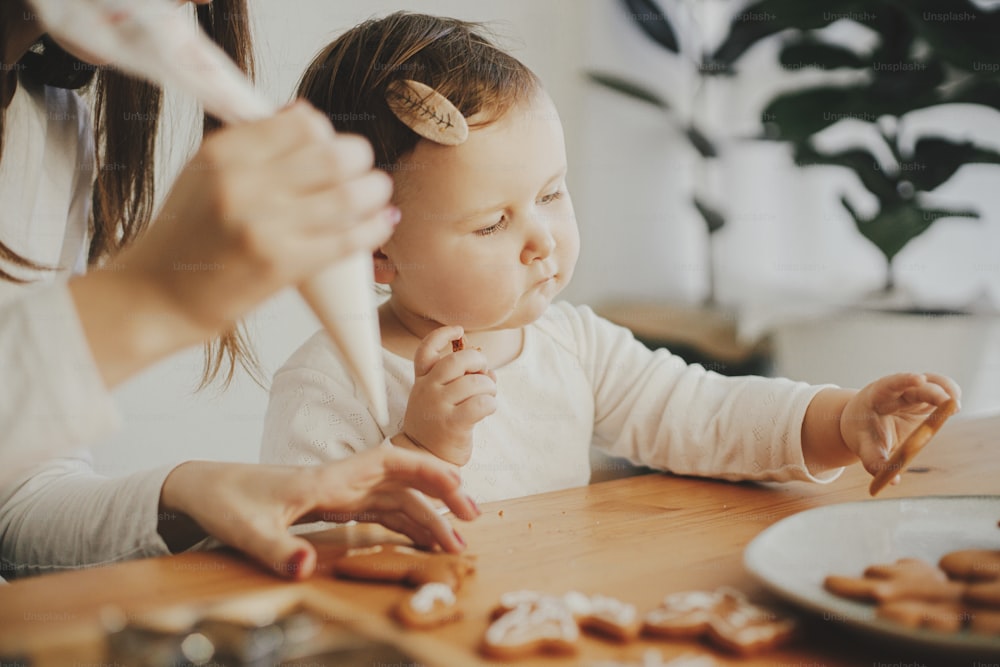 Cute little daughter and mother together decorating christmas gingerbread cookies with icing on wooden table in modern room.  Mommy daughter authentic lovely moments. Holiday preparations