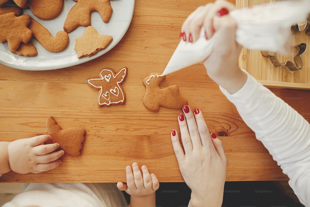 Cute little daughter and mother decorating christmas gingerbread cookies with icing on wooden table, authentic lovely moment. Family time together, holiday preparations. Top view