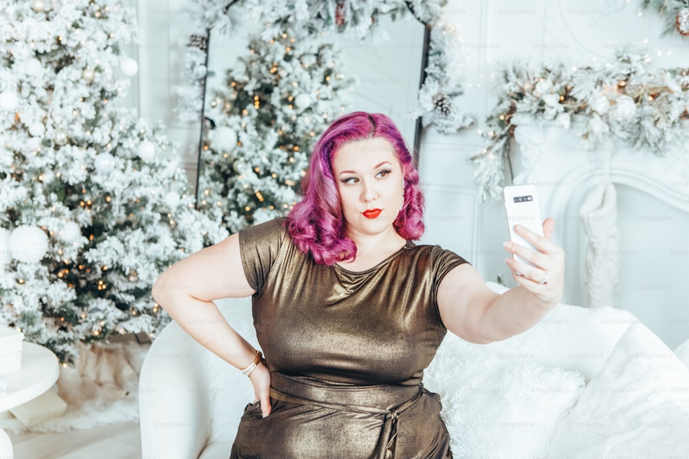 Hermosa joven caucásica modelo de talla grande mujer en vestido largo de color oliva dorado tomando selfie foto retrato en decoración de invierno de vacaciones en casa.