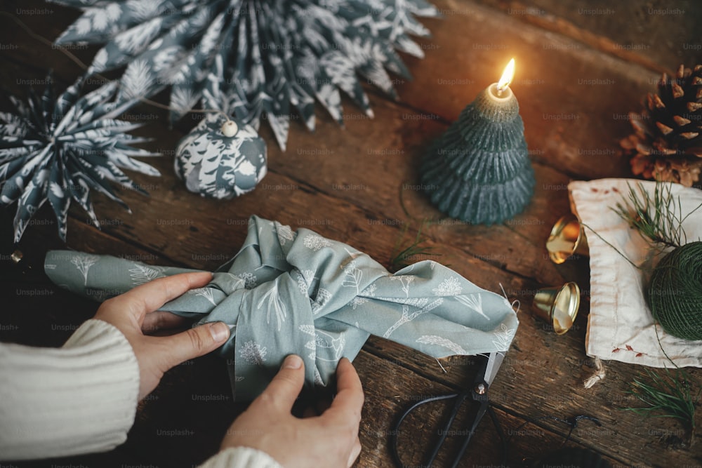 Furoshiki wrap. Hands wrapping christmas gift in fabric on rustic wooden table with candle, scissors, blue paper stars. Atmospheric moody image, nordic style. Merry Christmas and Happy Holidays!