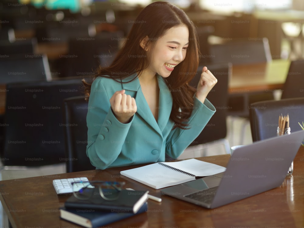 成功したビジネスウーマン、ノートパソコンを見ながら手を挙げて祝う女性社員。ワクワクして、とても幸せです。