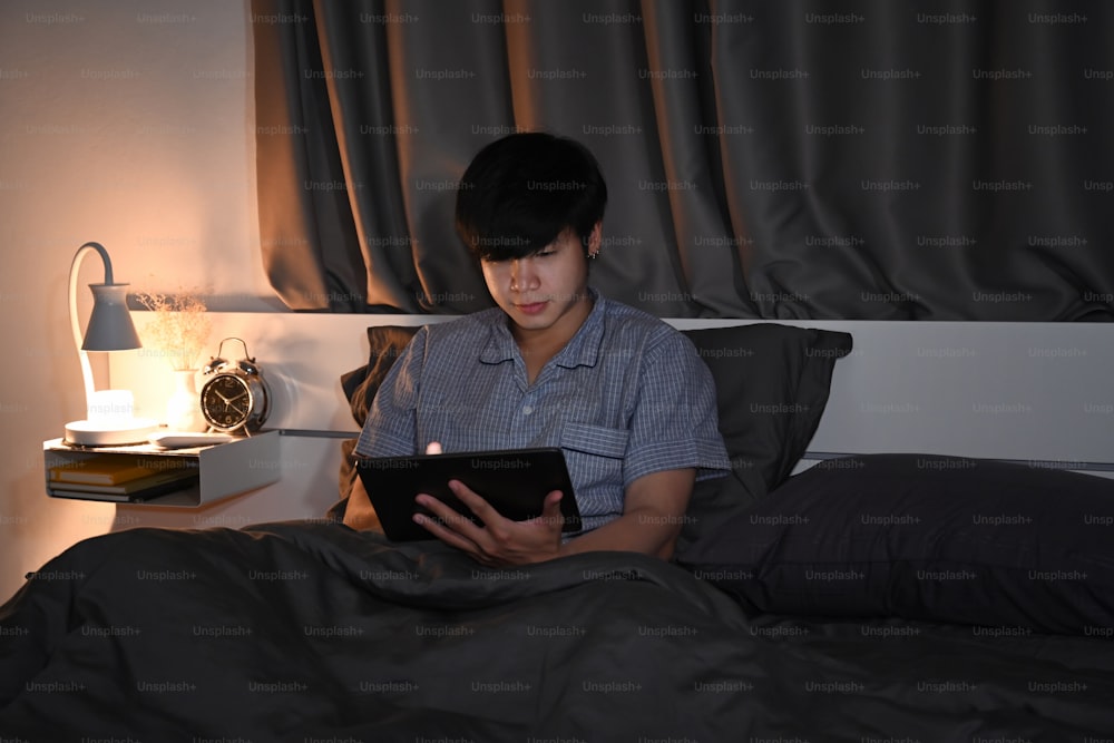 잠옷을 입은 청년이 침대에서 디지털 태블릿을 사용하고 있다.