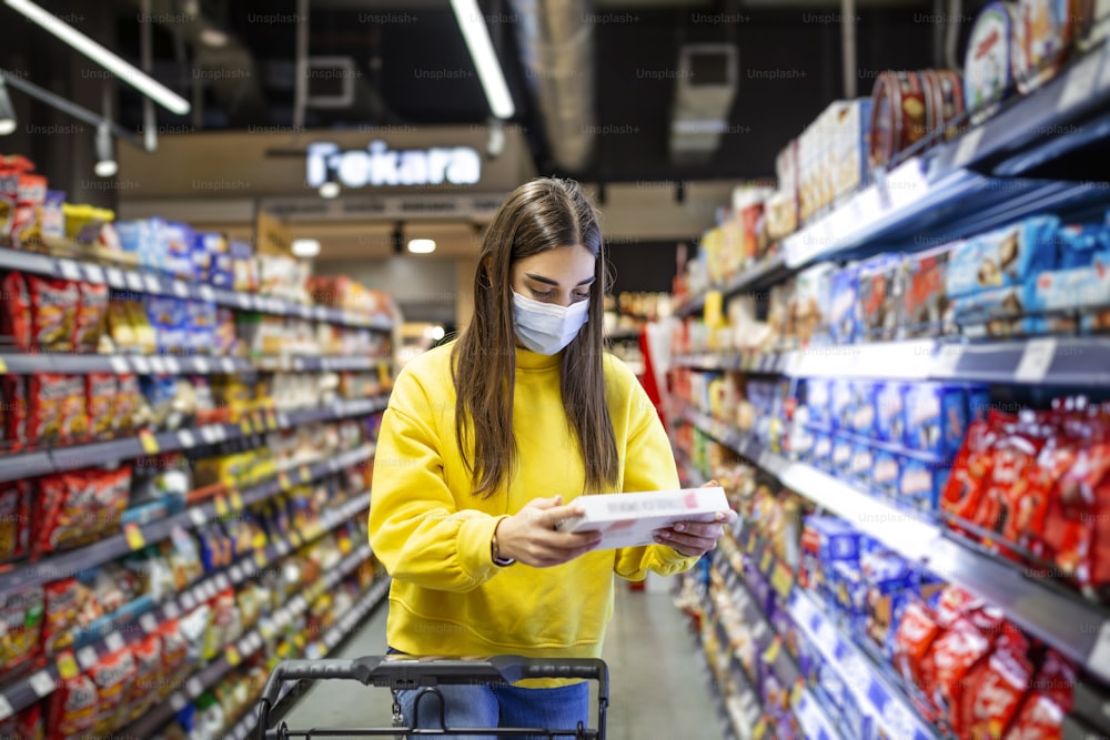 Social Distancing in einem Supermarkt. Eine junge Frau mit einer Einweg-Gesichtsmaske kauft Lebensmittel und legt sie in einen Einkaufskorb. Einkaufen während der Coronavirus-Covid-19-Epidemie