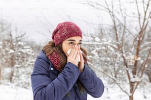 Young woman blowing in a tissue in a cold winter with a snowy mountain in the background. Portrait of a woman with tissue in hands looking away. Girl has the flu and running nose