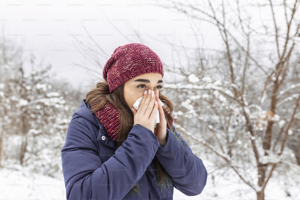 Young woman blowing in a tissue in a cold winter with a snowy mountain in the background. Portrait of a woman with tissue in hands looking away. Girl has the flu and running nose