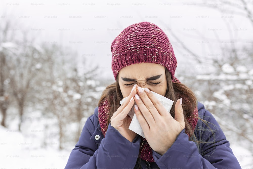 Mujer joven que sufre de un resfriado estacional y gripe se suena la nariz con un pañuelo mientras está al aire libre en invierno. Concepto médico y de atención médica