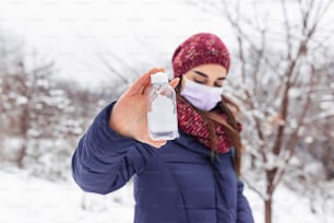 Young woman holding bottle of disinfectant. Girl wearing face mask. Woman showing how to stop spreading virus. Girl uses hand sanitizer outdoors
