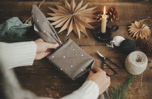 Hands wrapping christmas gift box in brown fabric on rustic wooden table with scissors, craft paper star, candle. Zero waste and eco friendly presents. Atmospheric moody time, nordic style.