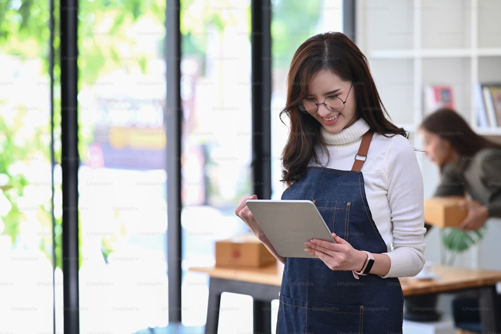 Cheerful young Asian woman entrepreneur using digital tablet and standing in her coffee shop.