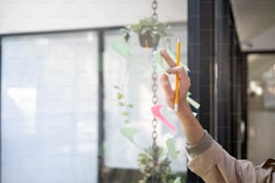Designer feminina de tiro cortado colocando nota adesiva colorida em uma parede de vidro em offie.