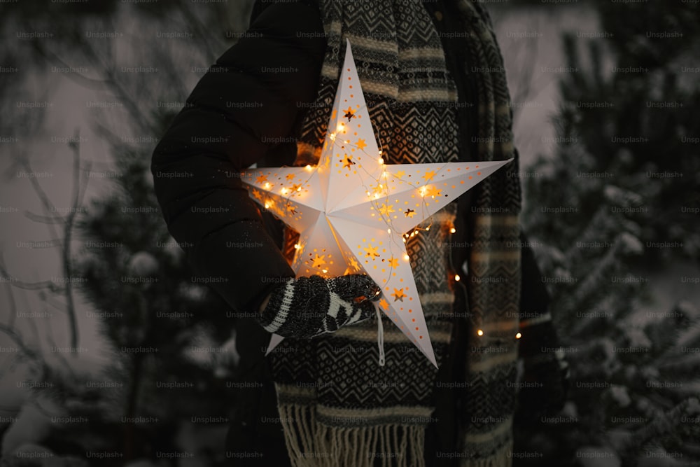 Woman holding big illuminated Christmas star in snowy winter park at night, close up. Magic Winter time. Stylish hipster female with glowing christmas star. Merry Christmas! Xmas miracle