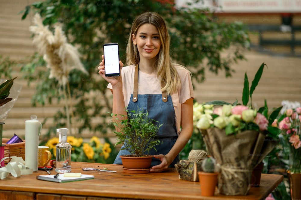 Woman florist working in a floral shop and showing a smartphone screen while taking care of a plant while standing next to the counter surrounded with flowers and plants