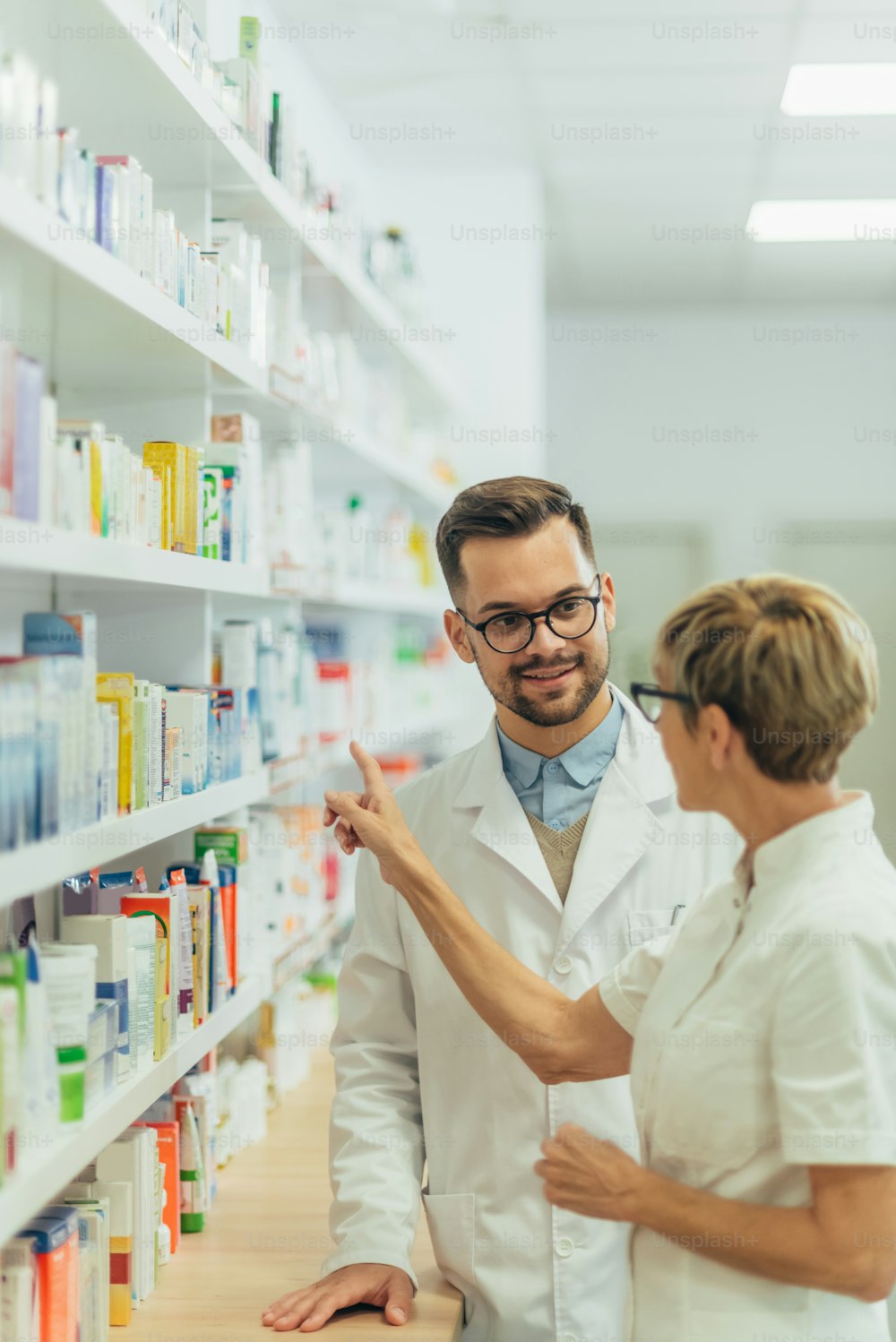 Happy young male pharmacist working with senior colleague while standing near the medicine shelf