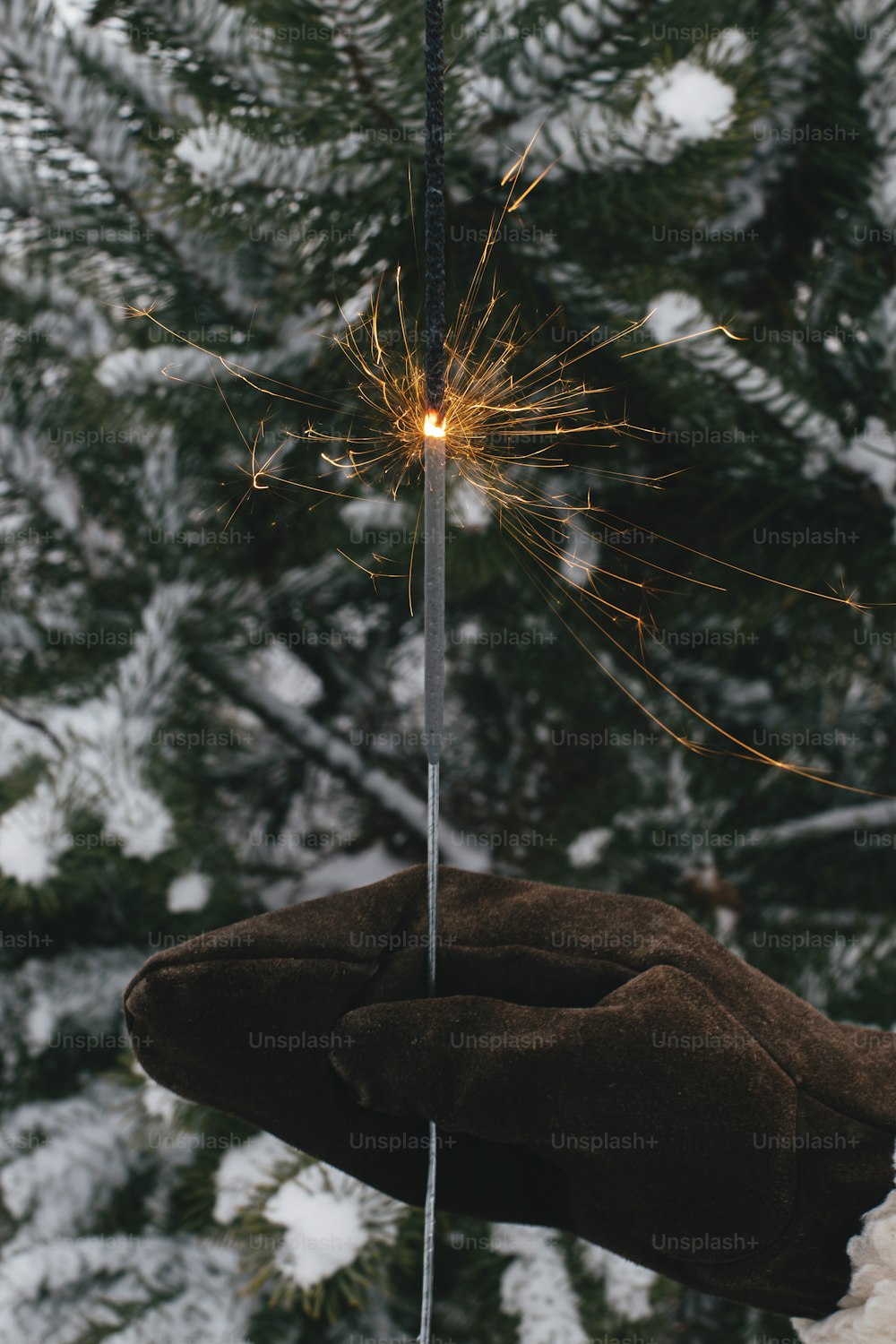 Happy New Year!  Woman hand in stylish mitten with glowing firework on background of pine tree branches in snow in evening. Hand holding burning sparkler. Atmospheric magic moment. Happy Holidays!
