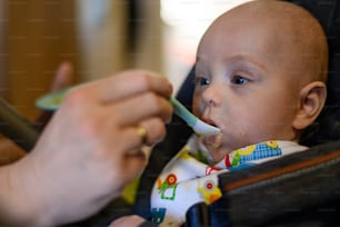 Portrait of an adorable cute baby eating with a face stained in food while mother feeds him with a spoon