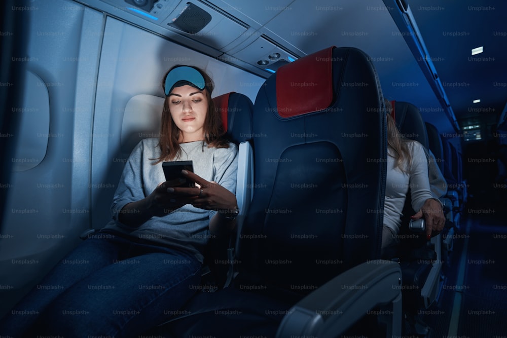 Woman traveler sitting in dim airplane cabin and using her mobile phone while having a flight