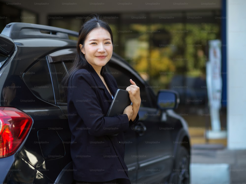 Smart asian businesswoman in black suit with digital tablet computer standing next to her car. Female car dealer.