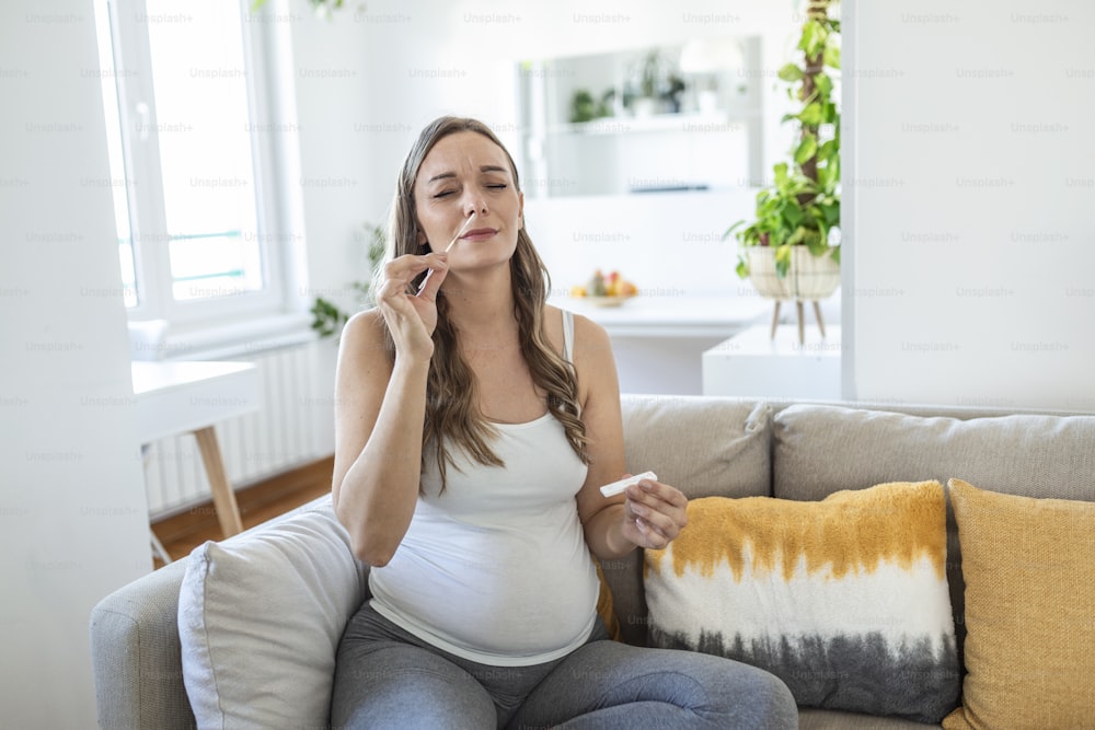 Pregnant woman sitting at a living room of a house with people, introducing a nasal swab in the nose to take an antigen coronavirus test, a Covid-19 rapid PCR to check sars infection.