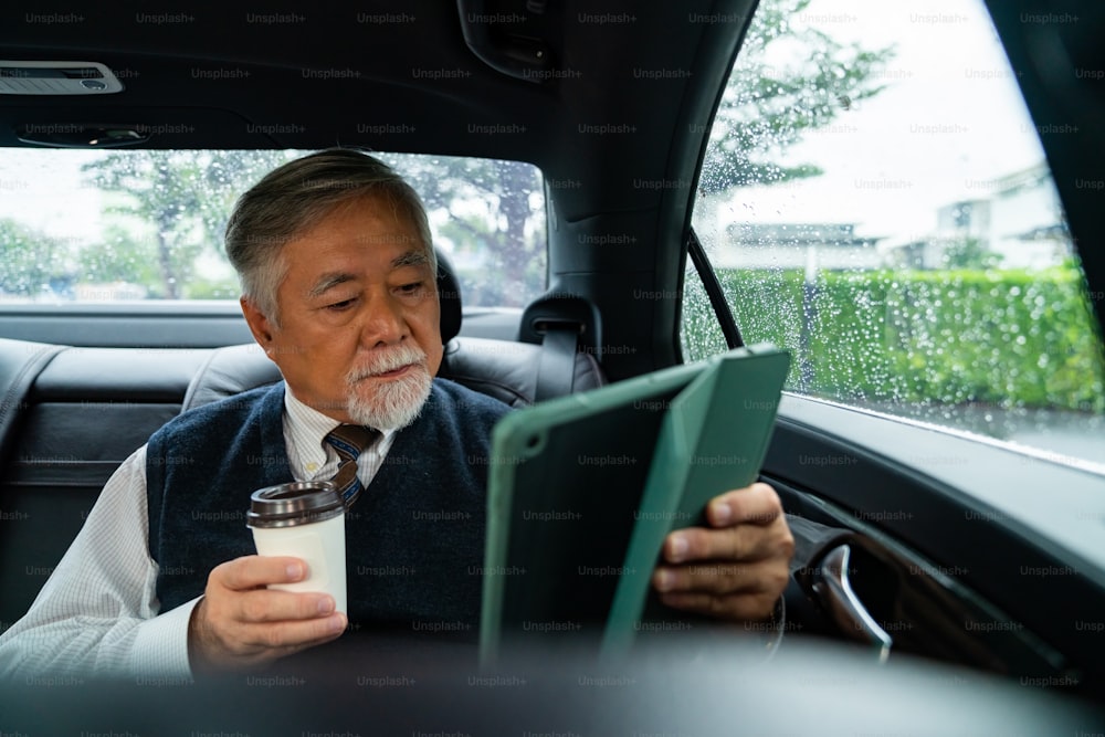 Confiance en soi homme d’affaires senior en costume assis sur la banquette arrière de la voiture buvant du café chaud tout en lisant le plan d’affaires sur une tablette numérique. PDG cadre âgé travaillant dans la conduite d’une voiture allant au bureau