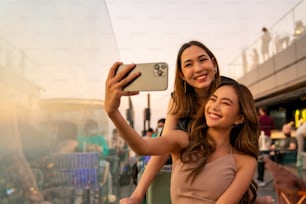 아시아 여자 친구들이 여름 해질녘 대도시의 마천루 옥상 레스토랑에서 만나 스마트폰 셀카를 함께 사용하고 있다. 여자 친구는 밤에 도시에서 야외 라이프 스타일 활동을 즐긴다