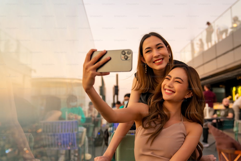 Des amies asiatiques se rencontrent et utilisent un selfie sur smartphone ensemble dans un restaurant sur le toit d��’un gratte-ciel dans une métropole au coucher du soleil d’été. Une amie profite d’une activité de plein air dans la ville la nuit