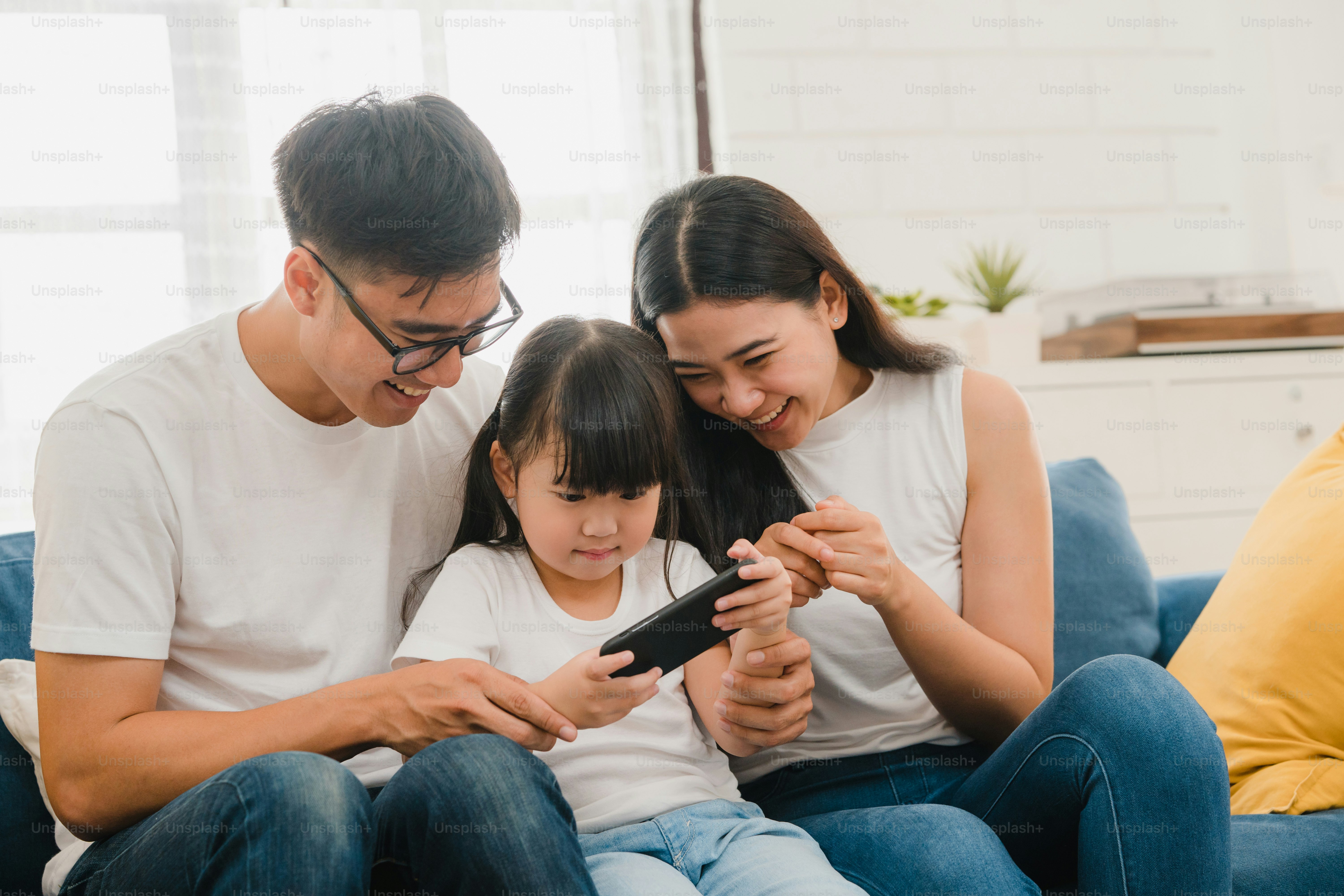 Happy Asian family dad, mom and daughter playing funny game online on smartphone sitting sofa in room at house.