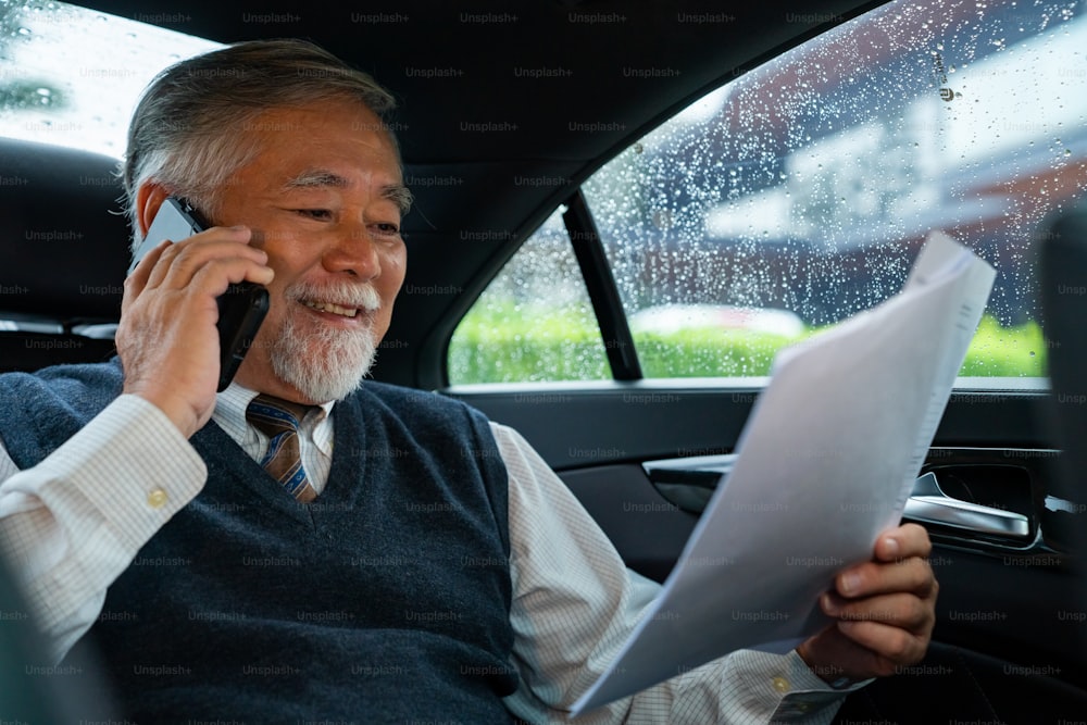 자신감 수석 사업가 CEO는 자동차 뒷좌석에 앉아 사무실에 가는 동안 휴대전화로 이야기하면서 사업 계획 문서를 읽고 있습니다. 노인 사업가와 교통 개념.