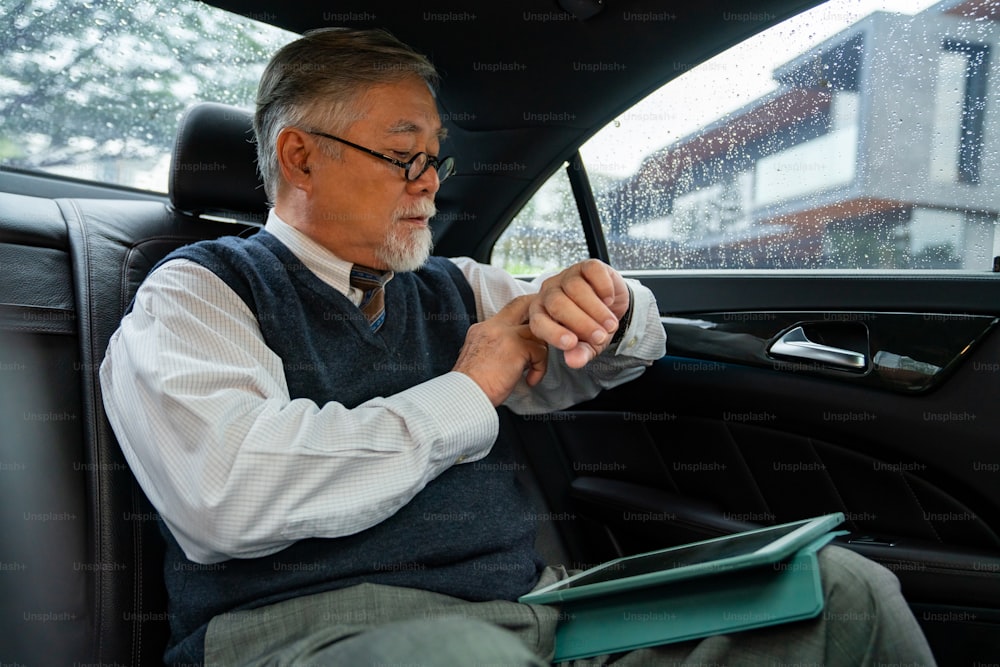 Confidence senior businessman CEO in suit sitting on car backseat working on digital tablet with checking time on hand watch while going to office. Elderly businessman and transportation concept.