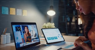 Una mujer asiática que usa una computadora portátil habla con colegas sobre el trabajo en videollamada mientras trabaja desde casa en la sala de estar por la noche.