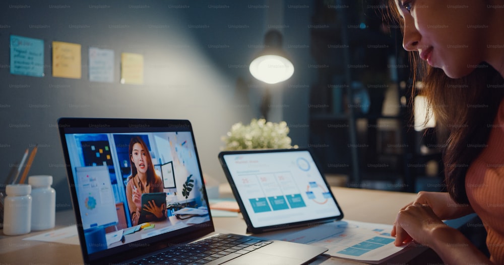 Asia woman using laptop talk to colleagues about work in video call while working from home at living room at night.