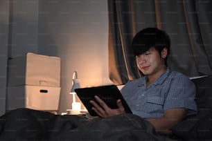 Smiling man sitting in bed at night and using digital tablet.