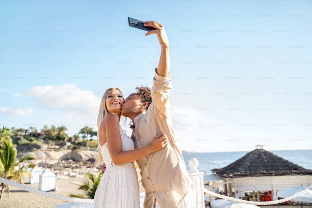 Happy couple in love kissing, taking selfie together with smartphone on the beach. Honeymoon. Travel. Vacation. Tourist. Playa del Duque. Tenerife.