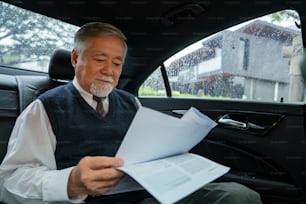 Hombre de negocios asiático de alto nivel con traje sentado en el asiento trasero del automóvil y escribiendo un plan de negocios mientras va a la oficina. Anciano CEO que trabaja en el automóvil y mira el tráfico por la ventana