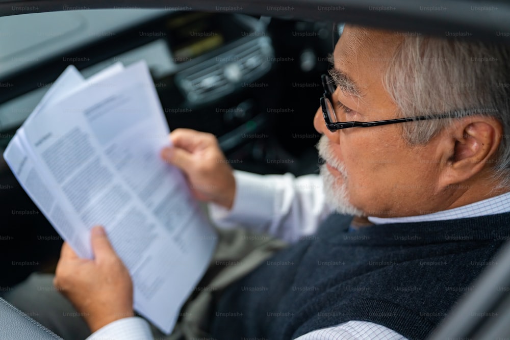 Confiança empresário sênior asiático de terno sentado no banco de trás do carro e escrevendo plano de negócios enquanto vai para o escritório. CEO idoso que trabalha em automóveis e olha para o tráfego pela janela