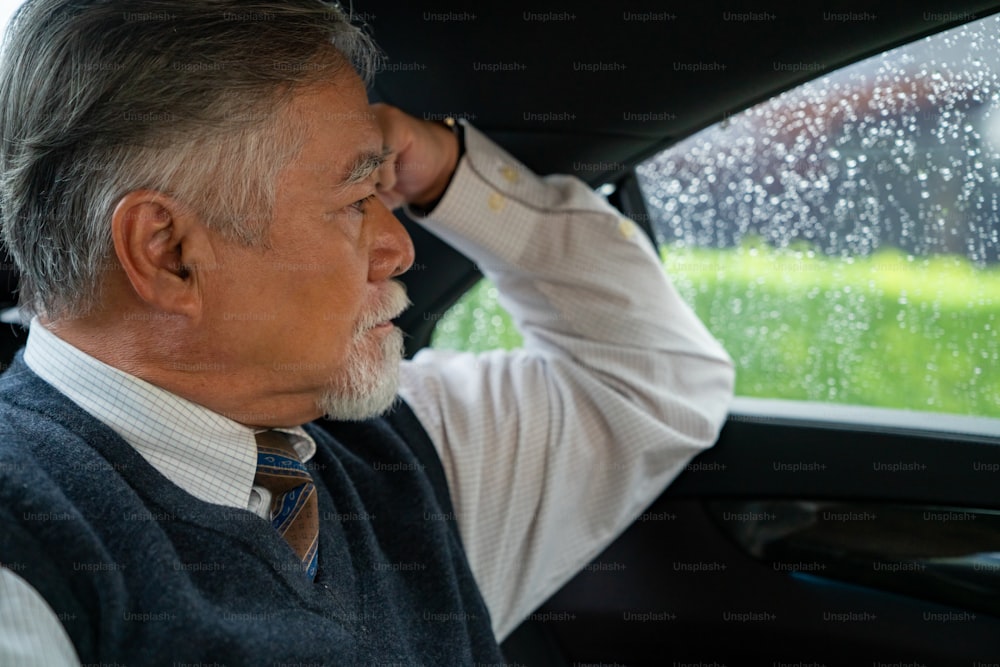 Confidence senior businessman in suit sitting on car backseat and thinking business plan while going to work at office. Elderly CEO working in automobile and looking at traffic out of the window