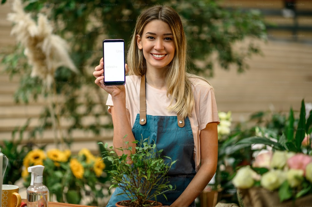 Woman florist working in a floral shop and showing a smartphone screen while taking care of a plant while standing next to the counter surrounded with flowers and plants