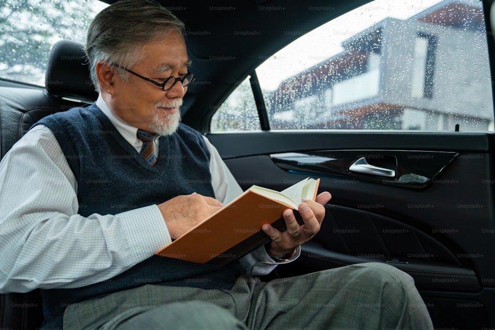 スーツを着た自信に満ちたシニアビジネスマンのCEOが眼鏡をかけ、車の後部座席に座り、オフィスで仕事に行きながら本を読んでいます。年配のビジネスマンと交通のコンセプト。