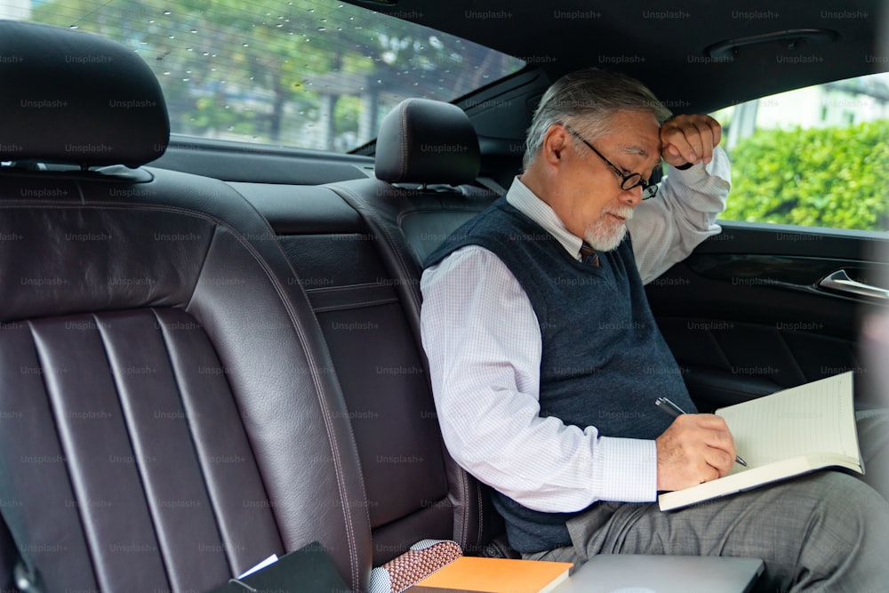 Vertrauen Senior Geschäftsmann CEO im Anzug mit Brille sitzt auf dem Rücksitz des Autos und schreibt Geschäftsplan in das Buch, während er zur Arbeit im Büro geht. Älterer Geschäftsmann und Transportkonzept.