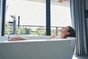 아침에 창가에 있는 욕조에서 졸고 있는 느슨한 검은 머리를 가진 차분한 편안한 여성
