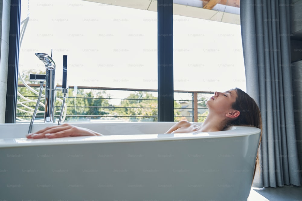 Calm relaxed female with loose dark hair dozing in bathtub by window in morning