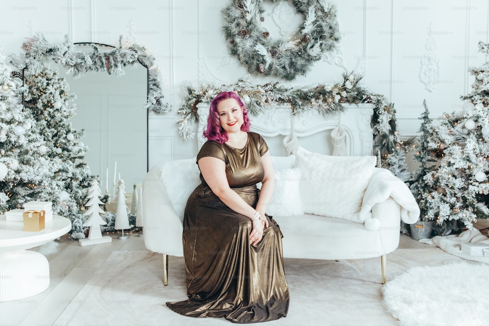 Beautiful young Caucasian plus size model woman in long golden olive color dress sitting on couch celebrating Christmas or New Year winter traditional holiday