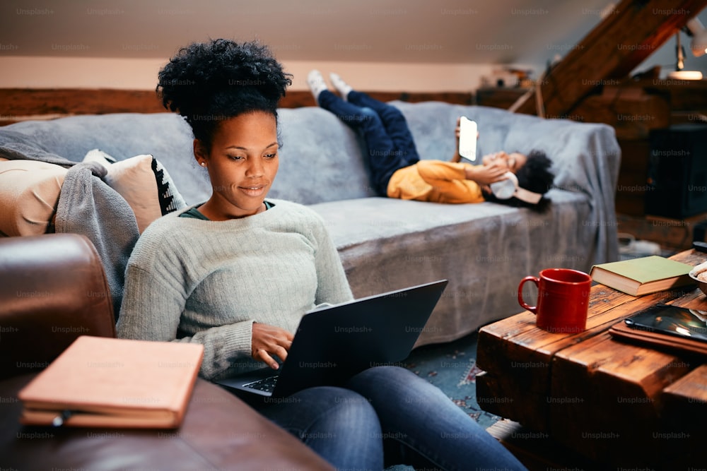 African American mother surfing the net on laptop while her daughter is relaxing on the sofa behind her.