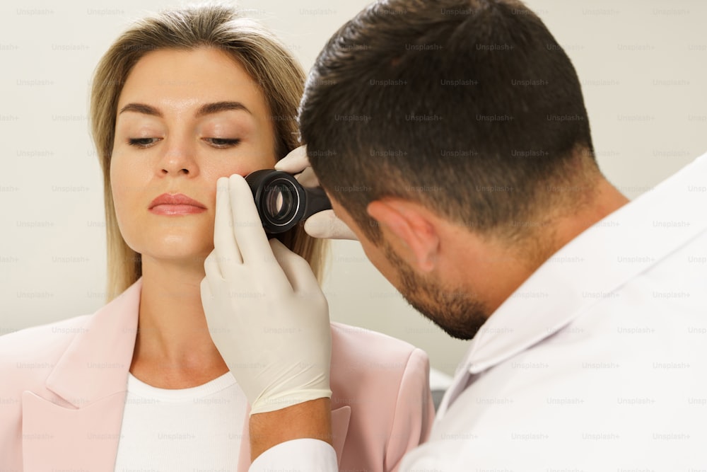 Médico y mujer cliente en clínica dermatológica. El dermatólogo está utilizando el dermatoscopio para el examen de la piel del rostro.
