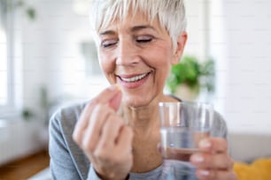 Smiling happy healthy middle aged 50s woman holding glass of water taking dietary supplement vitamin pill. Old women multivitamins antioxidants for anti age beauty.