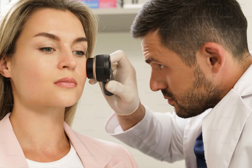 Doctor and client woman in dermatology clinic. Dermatologist is using dermatoscope for facial skin examination.