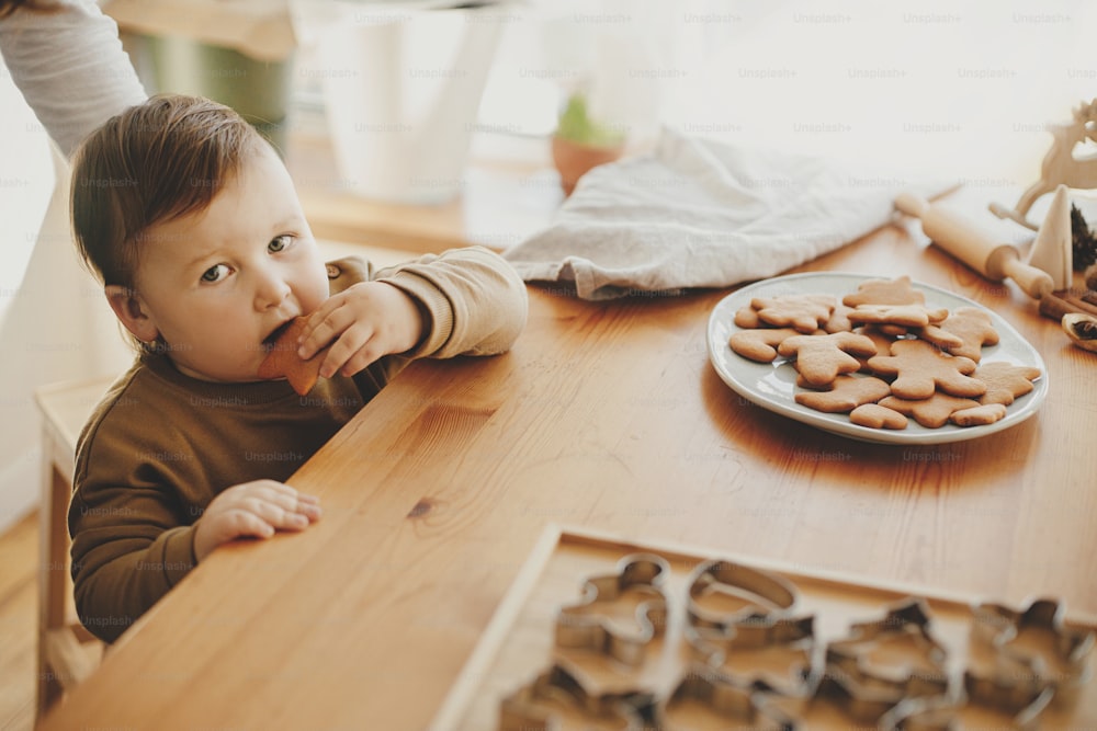 Adorable funny toddler tasting christmas cookies. Cute little girl eating freshly baked gingerbread cookie on wooden table in modern room. Authentic lovely moment, holiday preparations.