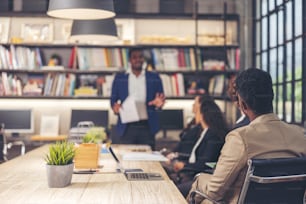 Multi-ethnic business team various office workers conversing and smiling while holding a meeting with colleagues in a modern office, senior businessman making a backdrop presentation