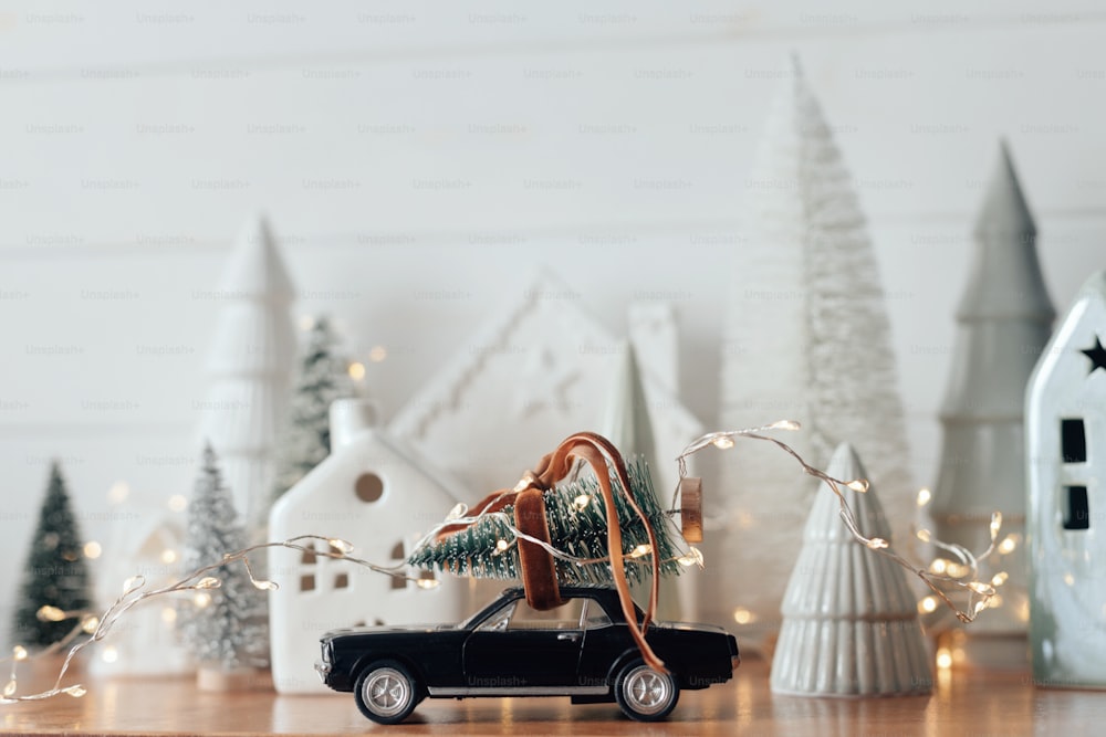 Les vacances arrivent ! Scène de Noël festive, village de vacances miniature. Voiture avec sapin de Noël, petites maisons et arbres enneigés sur fond blanc. Joyeux Noël et Bonne Année !